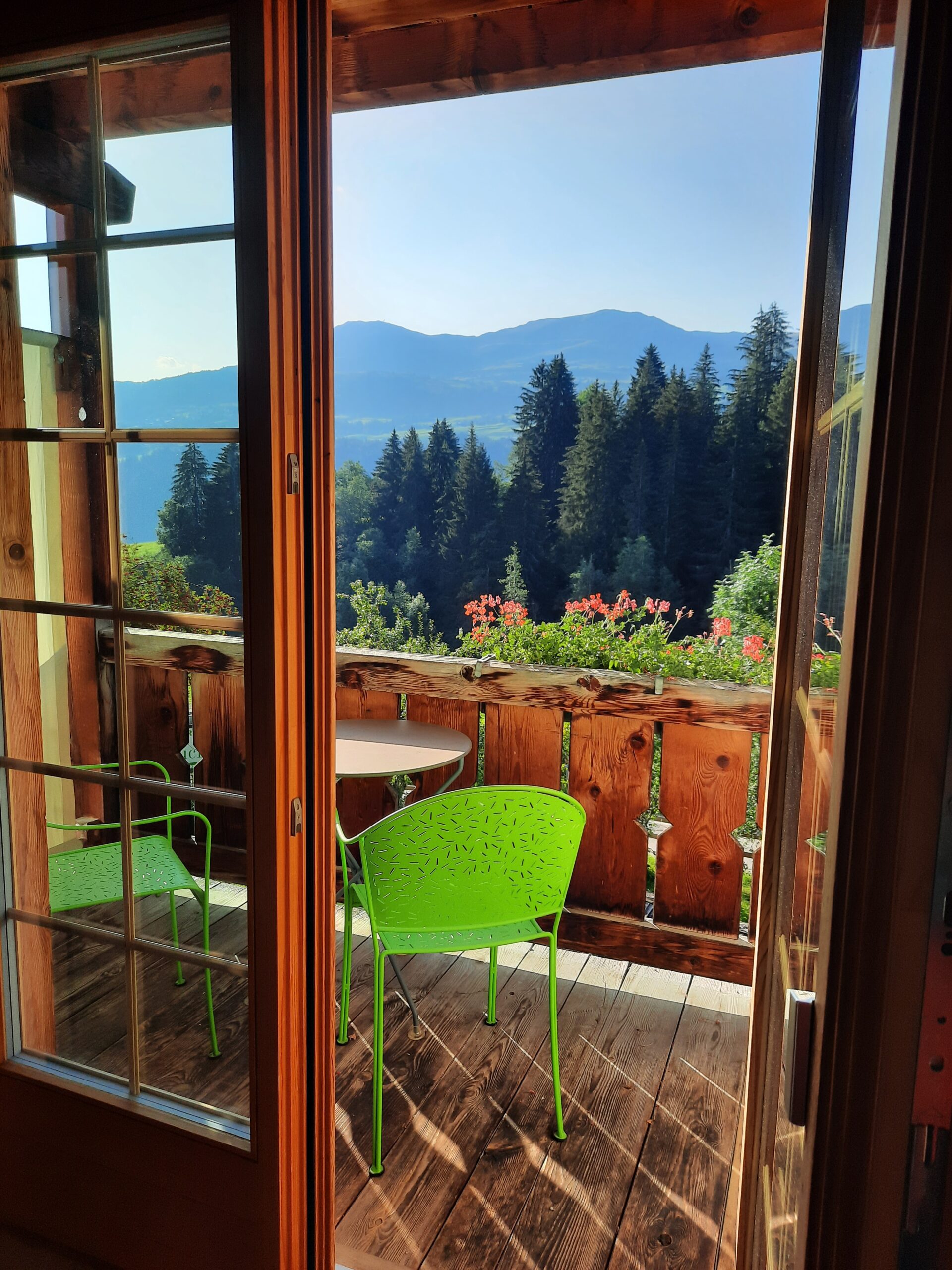 Blick vom Holzbalkon mit grünen Stühlen auf Berge und Wald.