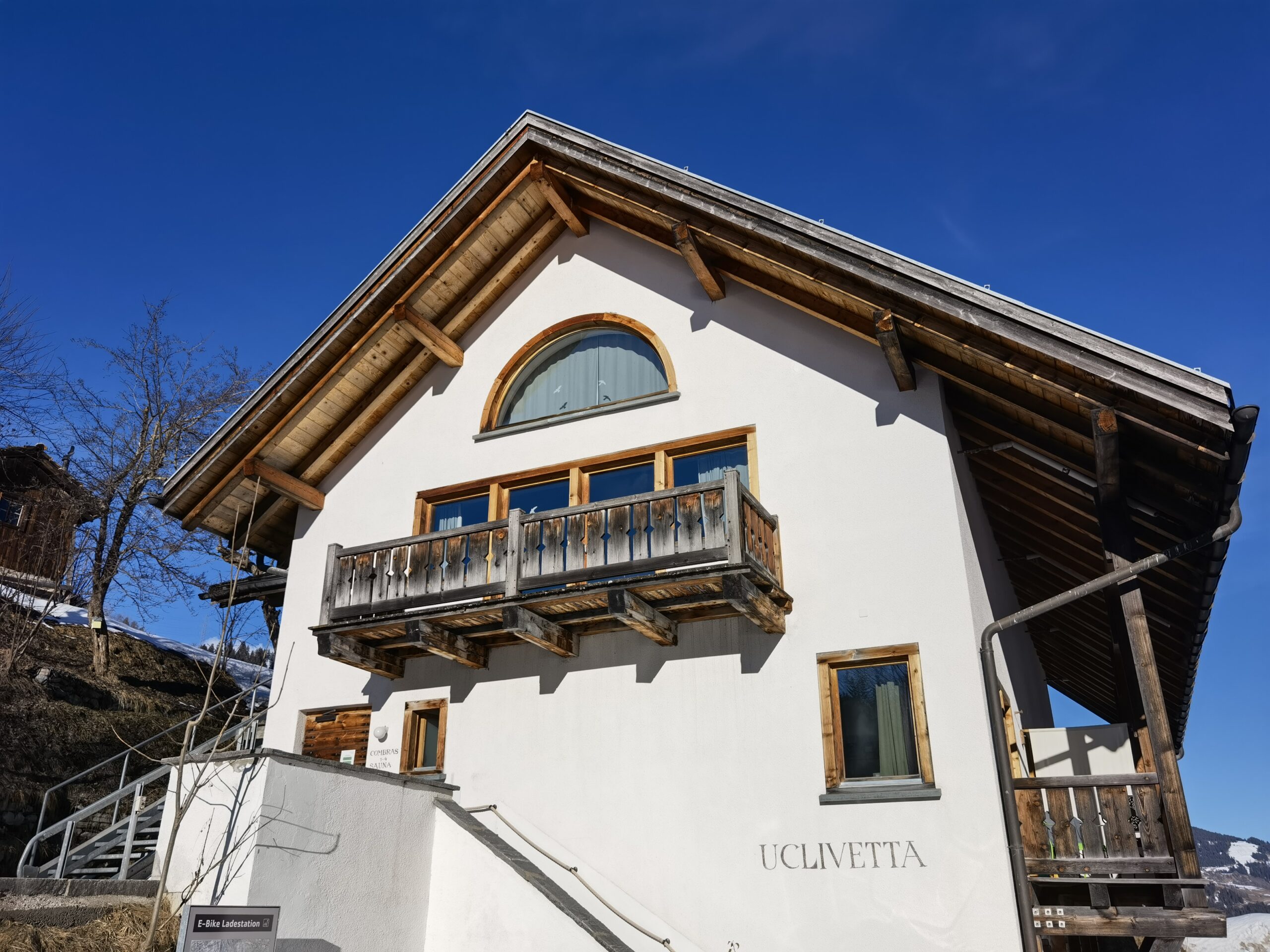Weißes Berghaus Uclivetta mit Holzbalkon unter blauem Himmel.